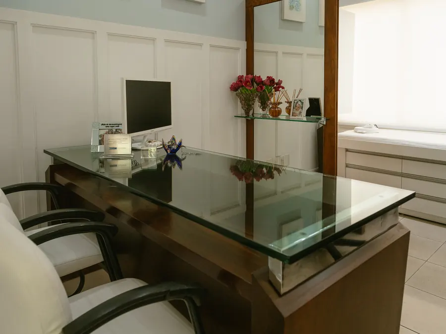 Mesa de vidro do consultório vista de frente, com poltronas e estante ao fundo
