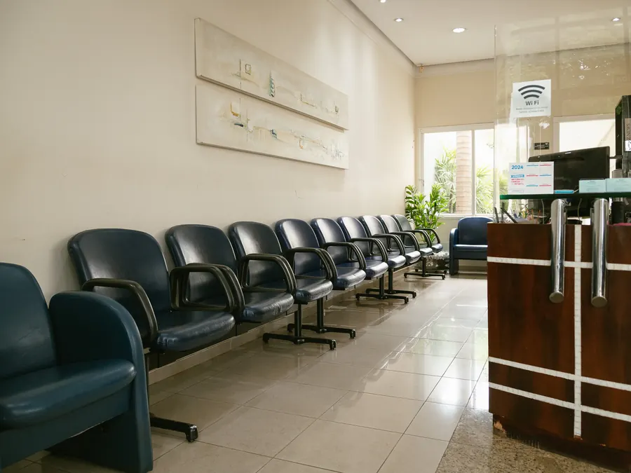 Sala de espera da clínica, com fileira de poltronas e o balcão da recepção ao fundo