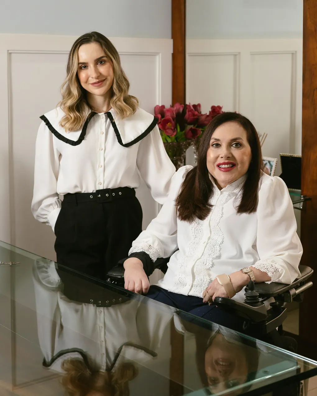 As duas dermatologistas à mesa de vidro do consultório: Dra. Fernanda em pé e Dra. Silvia Roberta sentada