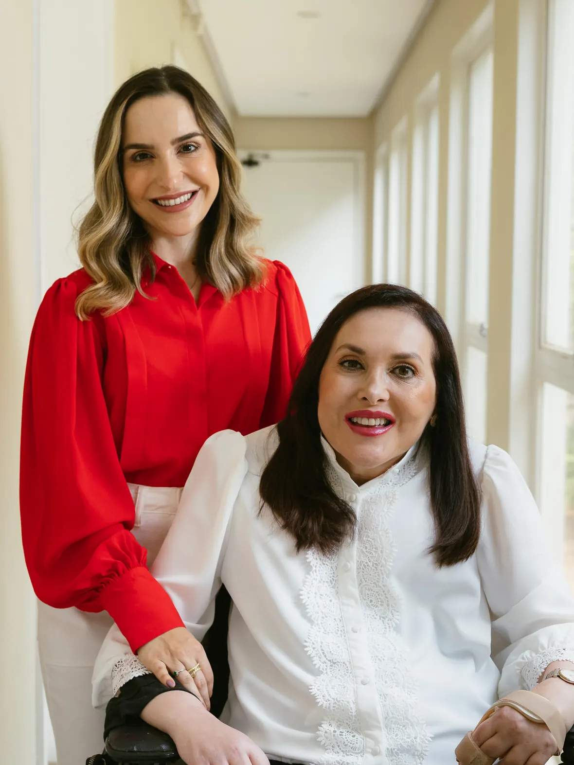 Dra. Fernanda Tirelli Rocha, de blusa vermelha, em pé atrás da Dra. Silvia Roberta Tirelli Rocha, sentada, no corredor iluminado da Clínica Renaissance